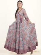A woman wearing a long, flowing dress with a detailed floral pattern in shades of red, blue, and white. The dress has three-quarter sleeves and a V-neckline, complemented by a tassel detail at the waist. She has long hair styled straight and is wearing earrings. The background is plain and light-colored.