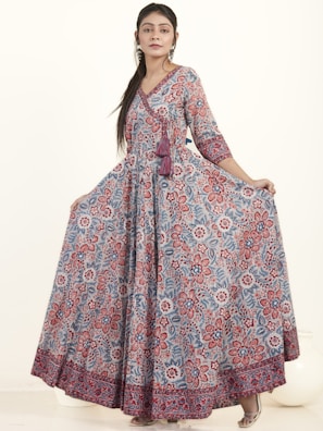A woman wearing a long, flowing dress with a detailed floral pattern in shades of red, blue, and white. The dress has three-quarter sleeves and a V-neckline, complemented by a tassel detail at the waist. She has long hair styled straight and is wearing earrings. The background is plain and light-colored.