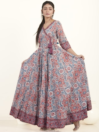 A woman wearing a long, flowing dress with a detailed floral pattern in shades of red, blue, and white. The dress has three-quarter sleeves and a V-neckline, complemented by a tassel detail at the waist. She has long hair styled straight and is wearing earrings. The background is plain and light-colored.