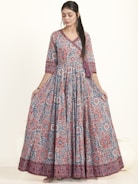 A woman is wearing an elegant floor-length dress adorned with a vibrant floral pattern in red and blue hues. The dress has a fitted top with long sleeves and a flowing skirt. She appears to be in a studio setting with a neutral background, showcasing the intricate design and bold colors of the outfit.