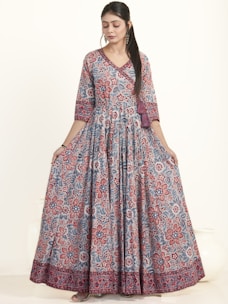 A woman is wearing an elegant floor-length dress adorned with a vibrant floral pattern in red and blue hues. The dress has a fitted top with long sleeves and a flowing skirt. She appears to be in a studio setting with a neutral background, showcasing the intricate design and bold colors of the outfit.