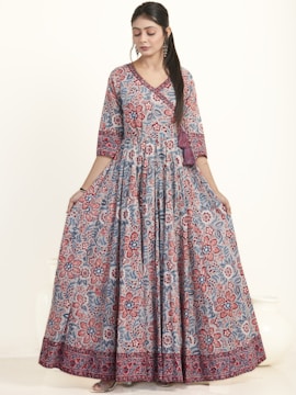 A woman is wearing an elegant floor-length dress adorned with a vibrant floral pattern in red and blue hues. The dress has a fitted top with long sleeves and a flowing skirt. She appears to be in a studio setting with a neutral background, showcasing the intricate design and bold colors of the outfit.