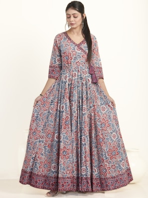 A woman is wearing an elegant floor-length dress adorned with a vibrant floral pattern in red and blue hues. The dress has a fitted top with long sleeves and a flowing skirt. She appears to be in a studio setting with a neutral background, showcasing the intricate design and bold colors of the outfit.