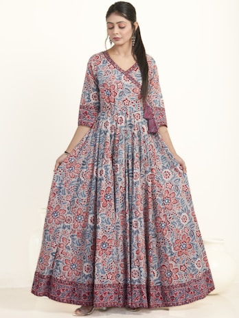 A woman is wearing an elegant floor-length dress adorned with a vibrant floral pattern in red and blue hues. The dress has a fitted top with long sleeves and a flowing skirt. She appears to be in a studio setting with a neutral background, showcasing the intricate design and bold colors of the outfit.