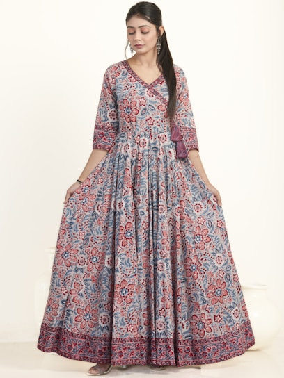 A woman is wearing an elegant floor-length dress adorned with a vibrant floral pattern in red and blue hues. The dress has a fitted top with long sleeves and a flowing skirt. She appears to be in a studio setting with a neutral background, showcasing the intricate design and bold colors of the outfit.