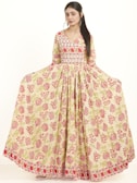 A woman is posing in a full-length, floral-patterned dress with long sleeves. The dress features a blend of pink and green floral designs on a cream-colored fabric, creating a graceful and elegant look.