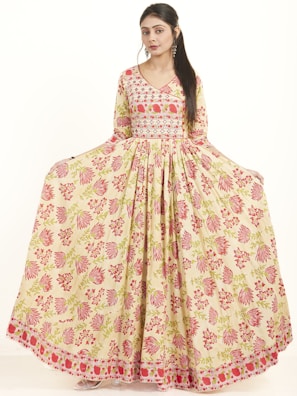 A woman is posing in a full-length, floral-patterned dress with long sleeves. The dress features a blend of pink and green floral designs on a cream-colored fabric, creating a graceful and elegant look.