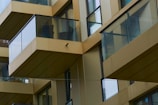 A series of modern glass and metal balconies on a high-rise building, reflecting a minimalist architectural style. The balconies have transparent glass railings and are staggered across different floors, creating a geometric pattern. The building's walls are clad in a light beige color, enhancing the sleek and contemporary design.