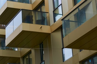 A series of modern glass and metal balconies on a high-rise building, reflecting a minimalist architectural style. The balconies have transparent glass railings and are staggered across different floors, creating a geometric pattern. The building's walls are clad in a light beige color, enhancing the sleek and contemporary design.