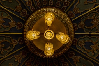 An ornate ceiling-mounted chandelier with five lit bulbs casts a warm glow. The chandelier features intricate golden designs and glass enclosures around the bulbs. The background is a richly decorated surface with artistic patterns and vivid colors.
