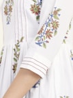 A close-up of a garment with intricate floral embroidery featuring red and green flowers with blue accents, sewn onto a white fabric. The sleeves have detailed stitching with fine red lines adding to the design.