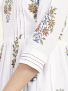 A close-up of a garment with intricate floral embroidery featuring red and green flowers with blue accents, sewn onto a white fabric. The sleeves have detailed stitching with fine red lines adding to the design.