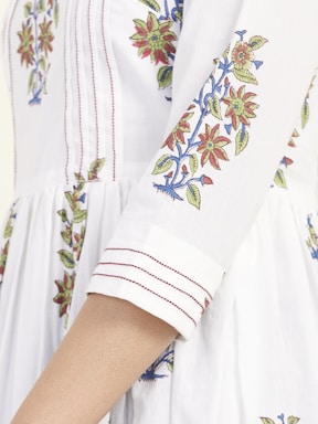 A close-up of a garment with intricate floral embroidery featuring red and green flowers with blue accents, sewn onto a white fabric. The sleeves have detailed stitching with fine red lines adding to the design.