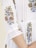 A close-up view of a white garment with intricate floral embroidery in red, green, and blue. The embroidery features detailed flowers and leaves running down the sleeves and along the seams. The garment has red stitched pleats and a relaxed fit, giving it an elegant appearance.