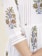 A close-up view of a white garment with intricate floral embroidery in red, green, and blue. The embroidery features detailed flowers and leaves running down the sleeves and along the seams. The garment has red stitched pleats and a relaxed fit, giving it an elegant appearance.