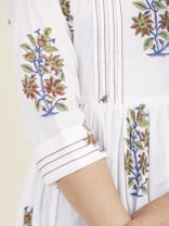 A close-up view of a white garment with intricate floral embroidery in red, green, and blue. The embroidery features detailed flowers and leaves running down the sleeves and along the seams. The garment has red stitched pleats and a relaxed fit, giving it an elegant appearance.