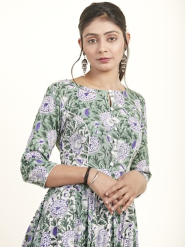 A person stands wearing a green floral dress with a detailed pattern of purple and white flowers. The dress has a buttoned front, and the person has long earrings and a braided hairstyle, with hands gently clasped in front.