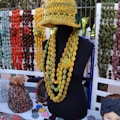 A display of handmade knitted items including a mannequin wearing a yellow-green knitted hat and several layered necklaces. Various colorful knitted scarves, hats, and accessories are arranged on a table and hanging in the background.
