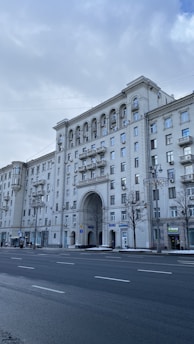 A large, classic-style apartment building with arches and decorative features is prominently displayed. The structure is adorned with multiple windows and balconies, and a wide street with some people walking alongside is visible in the foreground. Trees without leaves and overcast skies create a cold, urban atmosphere.