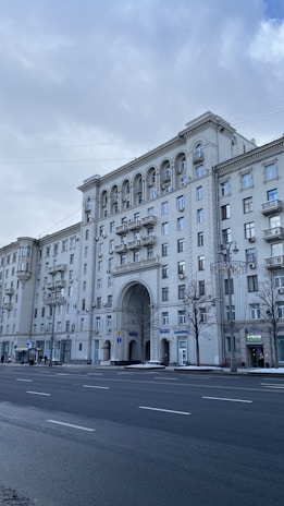 A large, classic-style apartment building with arches and decorative features is prominently displayed. The structure is adorned with multiple windows and balconies, and a wide street with some people walking alongside is visible in the foreground. Trees without leaves and overcast skies create a cold, urban atmosphere.