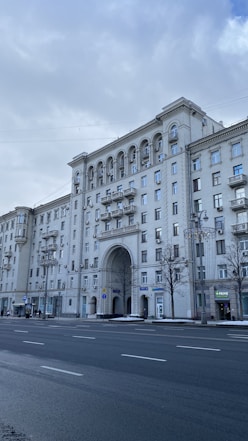A large, classic-style apartment building with arches and decorative features is prominently displayed. The structure is adorned with multiple windows and balconies, and a wide street with some people walking alongside is visible in the foreground. Trees without leaves and overcast skies create a cold, urban atmosphere.