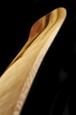 A close-up view of a wooden object with smooth curves and a polished surface. The grain of the wood is prominently displayed, showing natural tones ranging from light to medium brown. The background is stark black, highlighting the wood's texture and color.