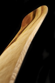 A close-up view of a wooden object with smooth curves and a polished surface. The grain of the wood is prominently displayed, showing natural tones ranging from light to medium brown. The background is stark black, highlighting the wood's texture and color.
