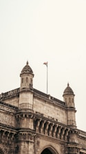 An ornate stone archway with intricate carvings, featuring two symmetrical towers topped with dome-like structures. A flagpole is mounted on the structure, flying a national flag against a light sky.