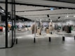 A modern clothing store with mannequins displaying beige and green outfits. The spacious, well-lit interior features neatly arranged racks of clothes and showcases a minimalist design. Spotlights hang from the ceiling, illuminating the display areas.