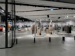 A modern clothing store with mannequins displaying beige and green outfits. The spacious, well-lit interior features neatly arranged racks of clothes and showcases a minimalist design. Spotlights hang from the ceiling, illuminating the display areas.