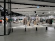 A modern clothing store with mannequins displaying beige and green outfits. The spacious, well-lit interior features neatly arranged racks of clothes and showcases a minimalist design. Spotlights hang from the ceiling, illuminating the display areas.