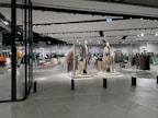 A modern clothing store with mannequins displaying beige and green outfits. The spacious, well-lit interior features neatly arranged racks of clothes and showcases a minimalist design. Spotlights hang from the ceiling, illuminating the display areas.