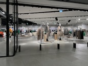 A modern clothing store with mannequins displaying beige and green outfits. The spacious, well-lit interior features neatly arranged racks of clothes and showcases a minimalist design. Spotlights hang from the ceiling, illuminating the display areas.