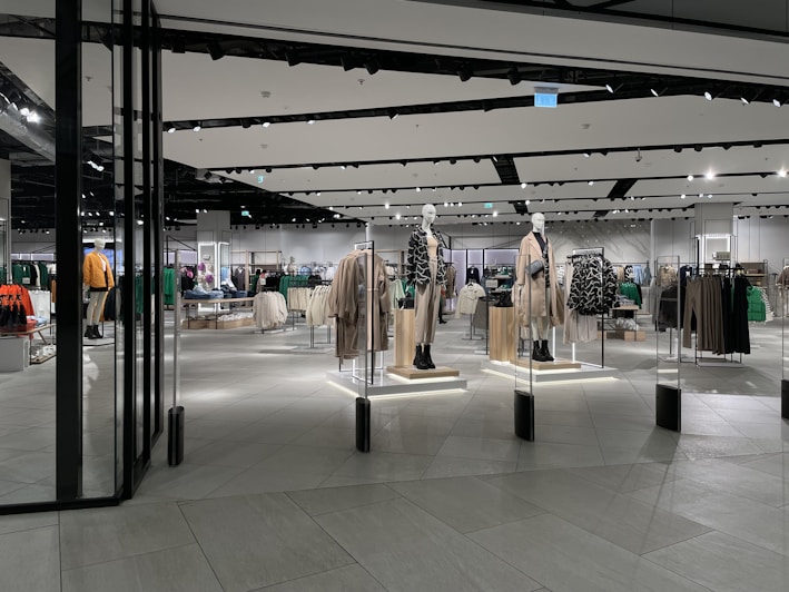 A modern clothing store with mannequins displaying beige and green outfits. The spacious, well-lit interior features neatly arranged racks of clothes and showcases a minimalist design. Spotlights hang from the ceiling, illuminating the display areas.