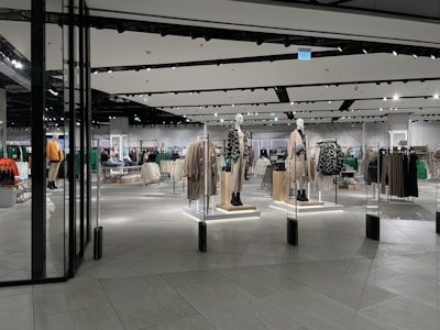 A modern clothing store with mannequins displaying beige and green outfits. The spacious, well-lit interior features neatly arranged racks of clothes and showcases a minimalist design. Spotlights hang from the ceiling, illuminating the display areas.