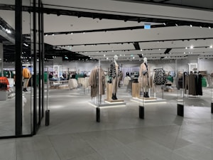 A modern clothing store with mannequins displaying beige and green outfits. The spacious, well-lit interior features neatly arranged racks of clothes and showcases a minimalist design. Spotlights hang from the ceiling, illuminating the display areas.