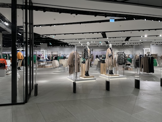 A modern clothing store with mannequins displaying beige and green outfits. The spacious, well-lit interior features neatly arranged racks of clothes and showcases a minimalist design. Spotlights hang from the ceiling, illuminating the display areas.
