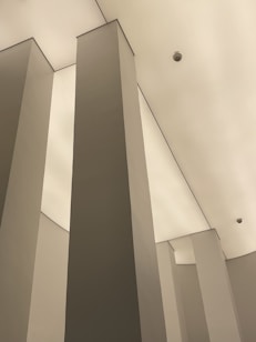 Tall, angular columns stretch upwards toward a smooth, pale ceiling. The minimalistic architectural design emphasizes vertical lines and geometric shapes. A surveillance camera is visible on the ceiling, enhancing the modern and secure ambiance.
