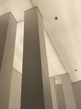 Tall, angular columns stretch upwards toward a smooth, pale ceiling. The minimalistic architectural design emphasizes vertical lines and geometric shapes. A surveillance camera is visible on the ceiling, enhancing the modern and secure ambiance.