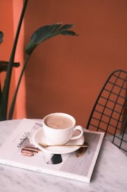A cup of coffee sits on a saucer atop a magazine titled 'cook life' on a marble table. A small spoon rests on the saucer beside the cup. In the background, there is a leafy green plant and an orange wall. A wireframe chair is partially visible on the right.