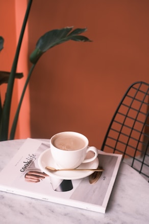 A cup of coffee sits on a saucer atop a magazine titled 'cook life' on a marble table. A small spoon rests on the saucer beside the cup. In the background, there is a leafy green plant and an orange wall. A wireframe chair is partially visible on the right.