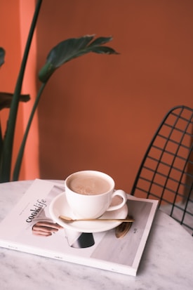 A cup of coffee sits on a saucer atop a magazine titled 'cook life' on a marble table. A small spoon rests on the saucer beside the cup. In the background, there is a leafy green plant and an orange wall. A wireframe chair is partially visible on the right.