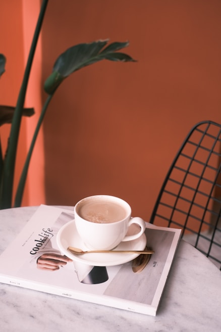 A cup of coffee sits on a saucer atop a magazine titled 'cook life' on a marble table. A small spoon rests on the saucer beside the cup. In the background, there is a leafy green plant and an orange wall. A wireframe chair is partially visible on the right.