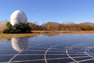 A white observatory dome is situated in a field with autumn-colored trees and a clear blue sky in the background. The foreground shows a reflective surface with grid markings that mirror the dome and landscape.