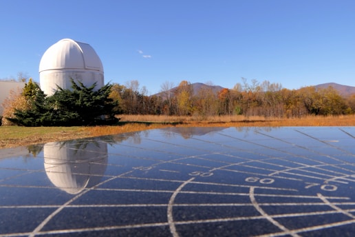 A white observatory dome is situated in a field with autumn-colored trees and a clear blue sky in the background. The foreground shows a reflective surface with grid markings that mirror the dome and landscape.