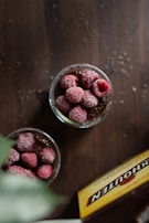 Several glass containers filled with chocolate chia seed pudding topped with frozen raspberries are arranged on a dark wooden table. A box with a partially visible brand name is nearby, and chia seeds are scattered on the surface.