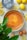 A freshly baked cheesecake is placed on a table surrounded by lemons, a container of Skyr yogurt, a knife with a yellow handle, and some eggs. A leafy green plant partially frames the image in the foreground.