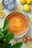 A freshly baked cheesecake is placed on a table surrounded by lemons, a container of Skyr yogurt, a knife with a yellow handle, and some eggs. A leafy green plant partially frames the image in the foreground.