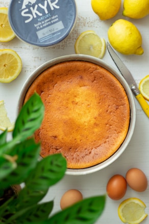 A freshly baked cheesecake is placed on a table surrounded by lemons, a container of Skyr yogurt, a knife with a yellow handle, and some eggs. A leafy green plant partially frames the image in the foreground.