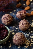 Round energy balls with bits of oats and dried fruit are displayed on a dark surface. A small bowl filled with dried cranberries is nearby, surrounded by scattered oats and almonds.